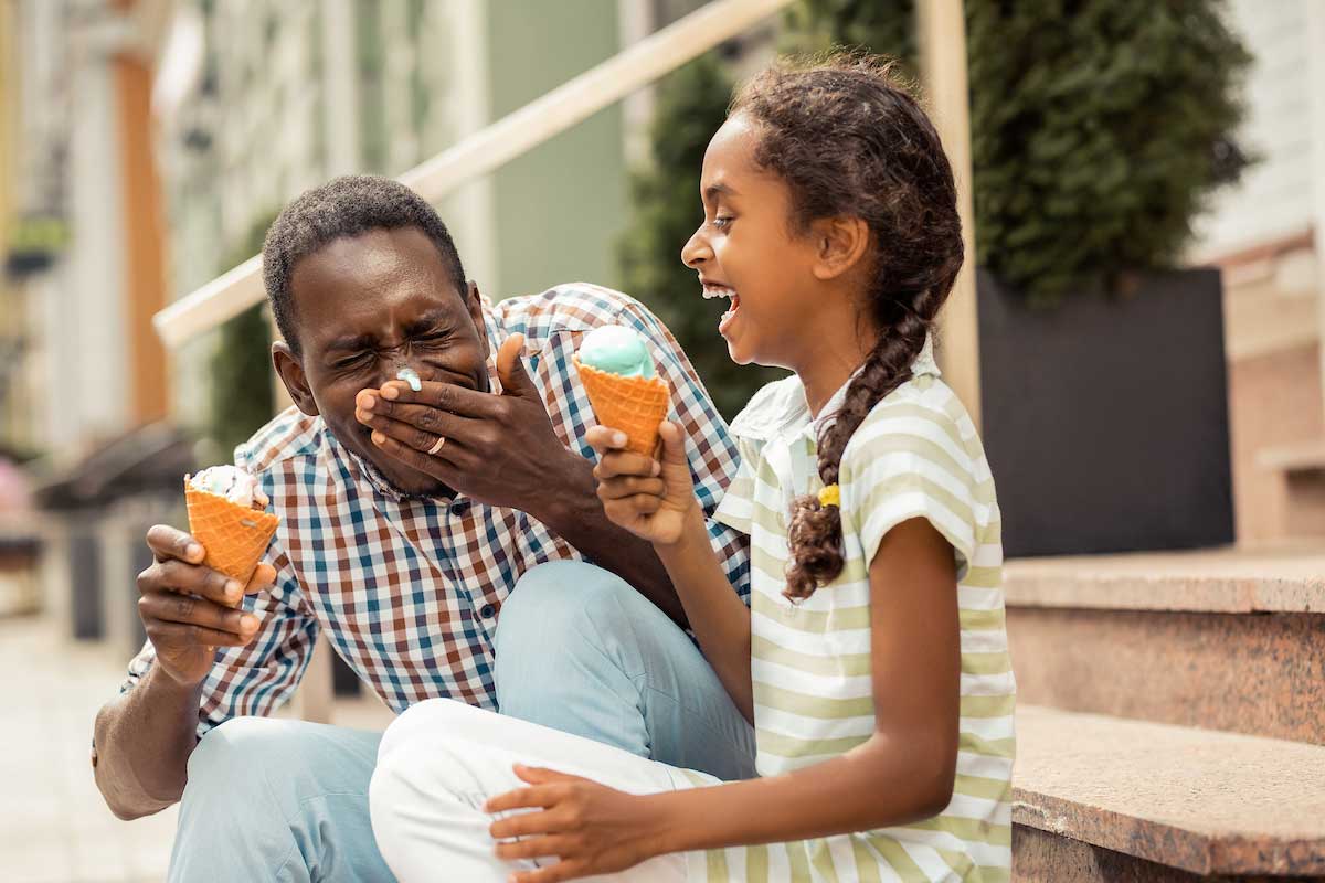 A father and daughter sit side by side outside on a set of stairs while laughing and enjoying ice cream cones together.