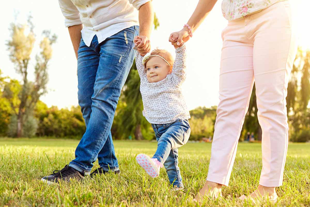 A baby is practicing walking outside in the grass with her parents holding each of her hands.